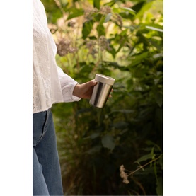 Model with Brabantia soft beige cup held outdoors, styled with white blouse and jeans, lush green foliage background