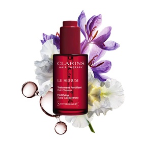 Clarins Le Serum Hair Serum bottle with dropper, surrounded by white and purple flowers and liquid droplets on white background