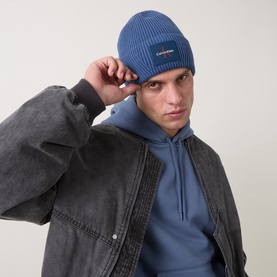Model with Calvin Klein Monogram Logo Patch Beanie Hat in Dark Denim, ribbed knit with turn-up brim, styled with grey jacket and blue hoodie