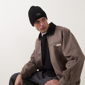 Model with Calvin Klein Monogram Logo Patch Beanie Hat in black, ribbed knit with turn-up brim, styled with a taupe harrington jacket