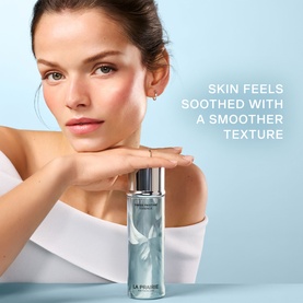 Model with La Prairie Swiss Pristine Essence 150ml toner, light blue frosted bottle with silver cap, resting chin on hands against pale blue