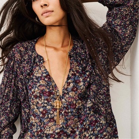 Model wearing Bash blue floral print V-neck blouse with long sleeves, styled with a gold tassel pendant necklace