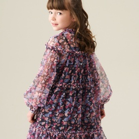 Model wearing a girls' purple floral chiffon dress with sheer puff sleeves, ruffle trim, and shirred bodice detail, seen from behind.