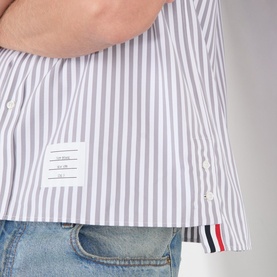 Model wearing Thom Browne medium grey stripe cotton casual shirt, with logo tab and tricolour flag detail at hem, styled with light blue denim jeans.