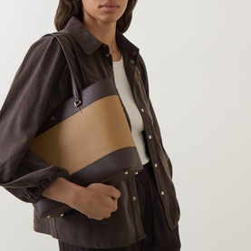 Model carrying Lauren Karly Logo Leather Tote Bag in camel and chestnut brown, with contrast stripe detail and metal monogram, styled with dark jacket