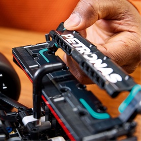 Hands assembling a black Lego Technic F1 car model, fitting a Petronas-branded component onto the detailed chassis build.