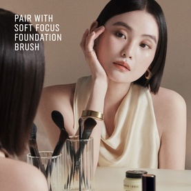 Model with makeup brushes in glass holder on vanity, wearing gold cuff bracelet and hoop earrings, soft neutral makeup look