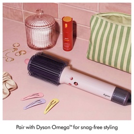 Dyson AirsmoothTM Styling Brush in pink and grey, shown with hair clips, gold rings, and a striped cosmetic bag on a pink surface.