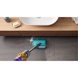 Dyson V15 Submarine vacuum in use on grey tiled floor near bathroom sink, emitting steam from teal wet cleaning head