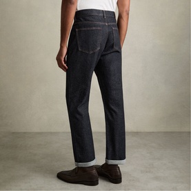 Model wearing Reiss raw indigo jeans, rear view, styled with a white tee and brown loafers, straight leg with cuffed hem.