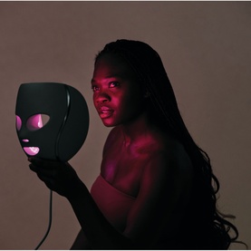 Model with Shark Beauty CryoGlow LED Face Mask Sage in black, held up showing pink LED light illumination through eye and mouth openings