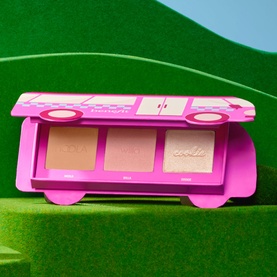 Benefit Make Up Set in pink bus-shaped palette containing three blush and highlight shades, displayed on green surface