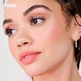 Model with Benefit makeup look featuring voluminous lashes, rosy blush, and a neutral lip, styled with gold bar earrings
