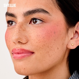 Model with Benefit make-up set; close-up showing rosy blush, freckles, and glossy pink lips on warm skin tone