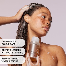 Model washing hair with Ouai clarifying shampoo, holding small white bottle, foam lather visible on hair and shoulder