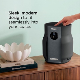Shark air purifier in dark grey with digital display, shown on wooden sideboard beside decorative bowl and books in a modern living space.