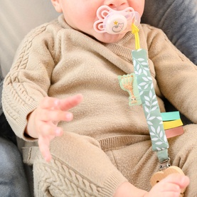 Baby holding Sophie the Giraffe multi-coloured dummy clip with pink soother attached, wearing a beige knit outfit
