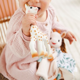Baby chewing Sophie the Giraffe teething toy in cream and gold spots, with matching soft giraffe toy visible in background