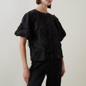 Model wearing FeeG black polyester top with floral sequin embellishment and puff sleeves, styled with black trousers