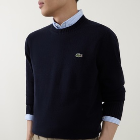 Model wearing Lacoste navy blue wool crew neck jumper with signature crocodile logo, styled over a light blue collared shirt