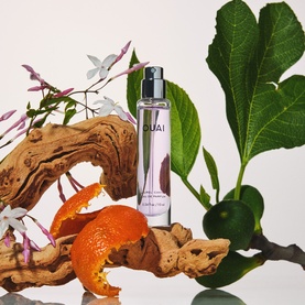 Ouai EDP fragrance spray bottle styled with driftwood, orange peel, white flowers and fig leaves on white background