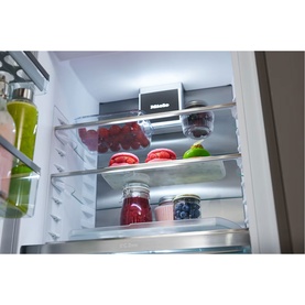 Miele Built-in 70/30 Fridge-Freezer interior showing illuminated glass shelves stocked with fresh fruit, vegetables and sealed food containers.