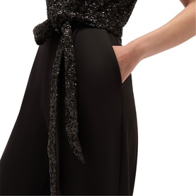 Model wearing Hobbs black dress with sequin embellished bodice and tie waist detail, hands in pockets of wide-leg skirt.