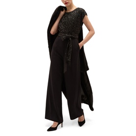Model wearing Hobbs black sequin embellished jumpsuit with wide-leg trousers, self-tie belt, and draped cape detail, styled with black heels.