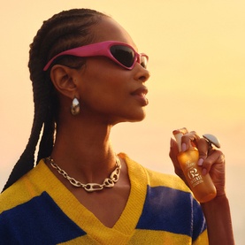 Model with Sol De Janeiro Cheirosa 62 Intense Perfume Mist, styled with pink sunglasses, chain necklace and striped knit top.