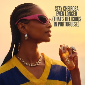 Model with Sol De Janeiro Cheirosa 62 Intense Perfume Mist, wearing pink sunglasses and chain necklace, yellow and blue striped top