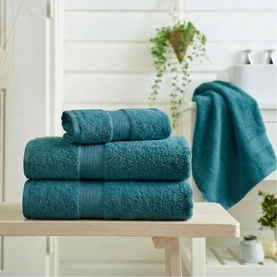 Deyongs Plain Towels in Topaz, a deep teal, shown as a stacked set of four on a wooden bench in a bathroom setting.