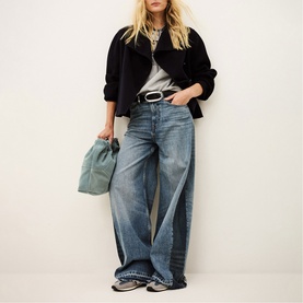 Model wearing Bash blue wide-leg jeans with black jacket, grey tee and belt, carrying a green tote bag.