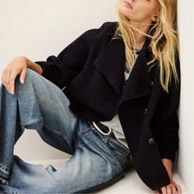 Model wearing Bash blue wide-leg jeans, styled with a black cropped jacket and grey top, seated against a white wall.