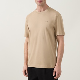 Model wearing Lacoste Crew Neck T-Shirt in beige cotton, featuring small logo embroidery on chest, paired with black trousers.