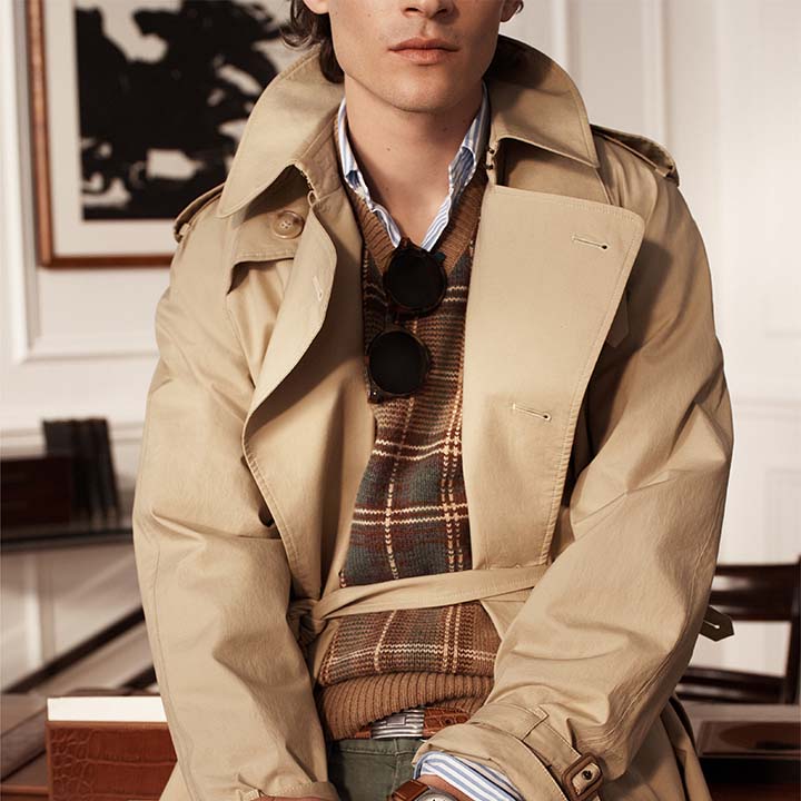 Person in a beige trench coat over a plaid sweater, wearing sunglasses, poses casually indoors.