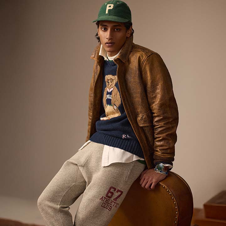 A person wearing a brown leather jacket, graphic sweater, and sweatpants, with a green cap, poses against a brown background.