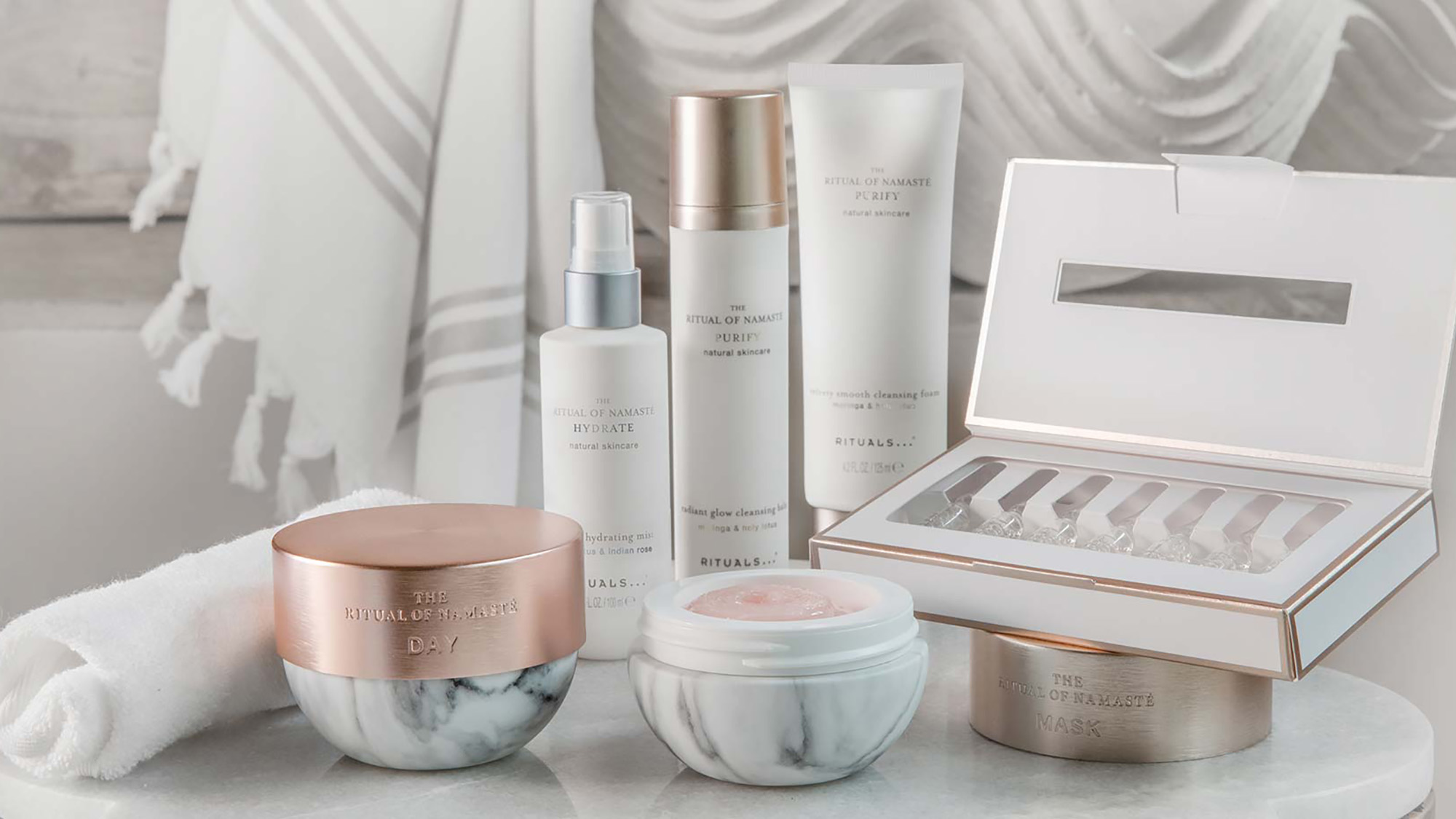 Collection of The Ritual of Namasté skincare products including creams, serums, cleansers and treatment masks in white and rose gold packaging.