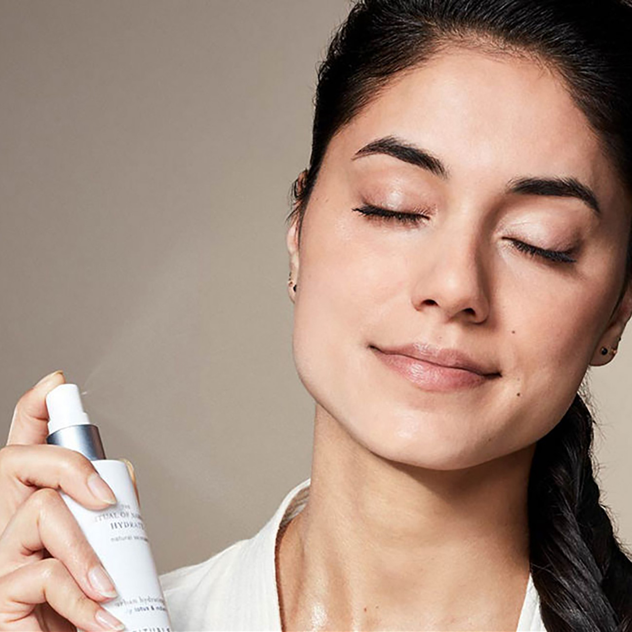 Woman applying hydrating mist spray to face, eyes closed, holding The Ritual of Namasté Hydrate skincare product.