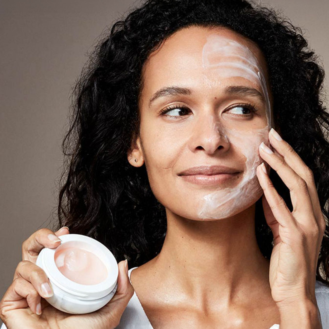 Woman applying face mask to cheeks, holding The Ritual of Namasté Glow product jar, showing skin brightening treatment.