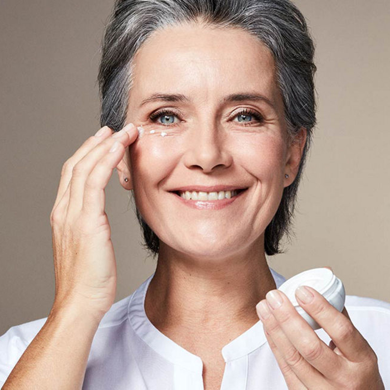 Woman smiling whilst applying eye cream, holding The Ritual of Namasté Ageless jar, demonstrating firming skincare.
