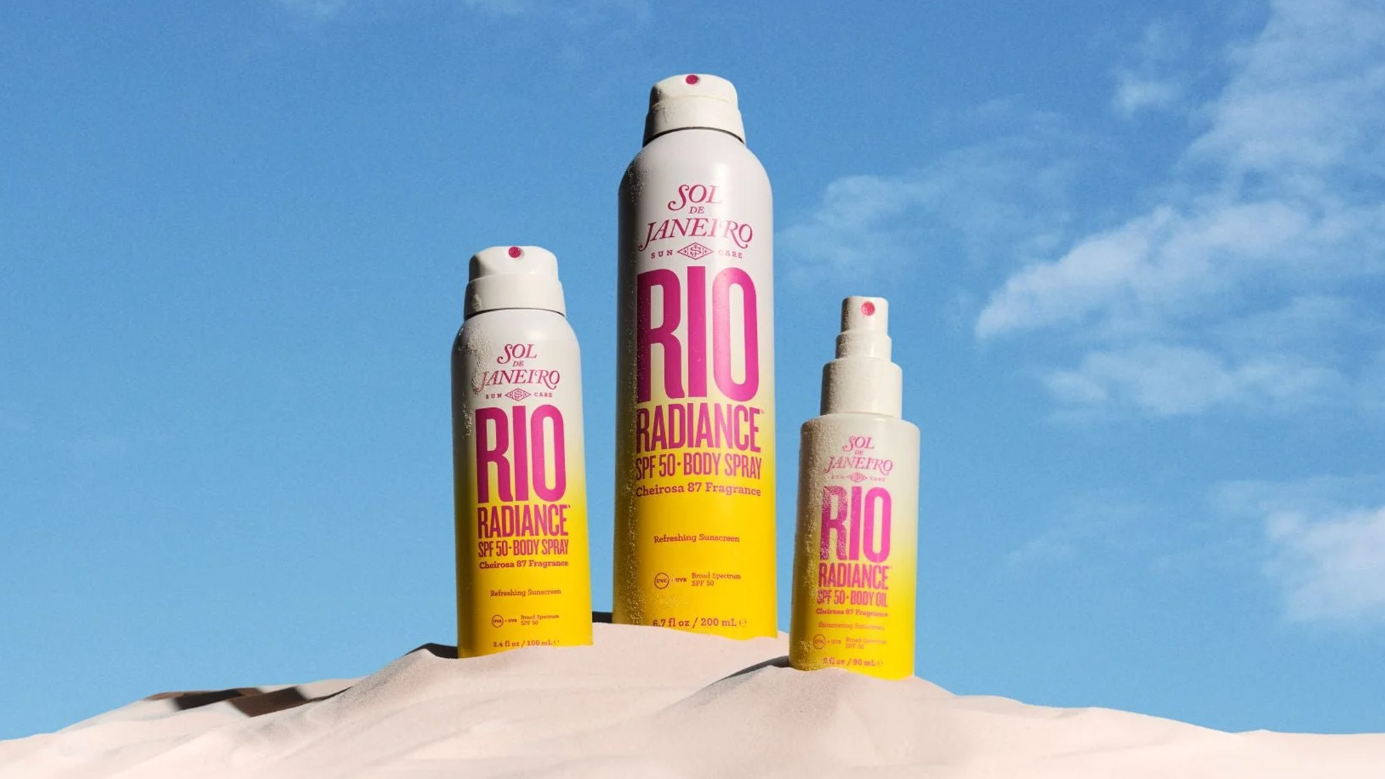 Three cans of Sol de Janeiro Rio Radiance SPF 30 body spray are displayed on sand against a blue sky.