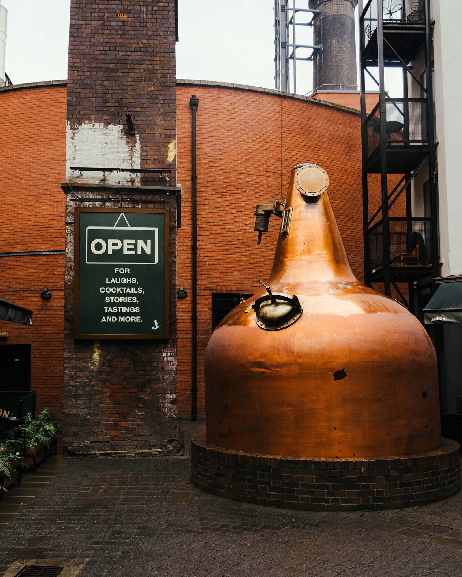 Copper distillery pot still outside historic brick building with green sign reading Open for laughs, cocktails, stories, tastings and more