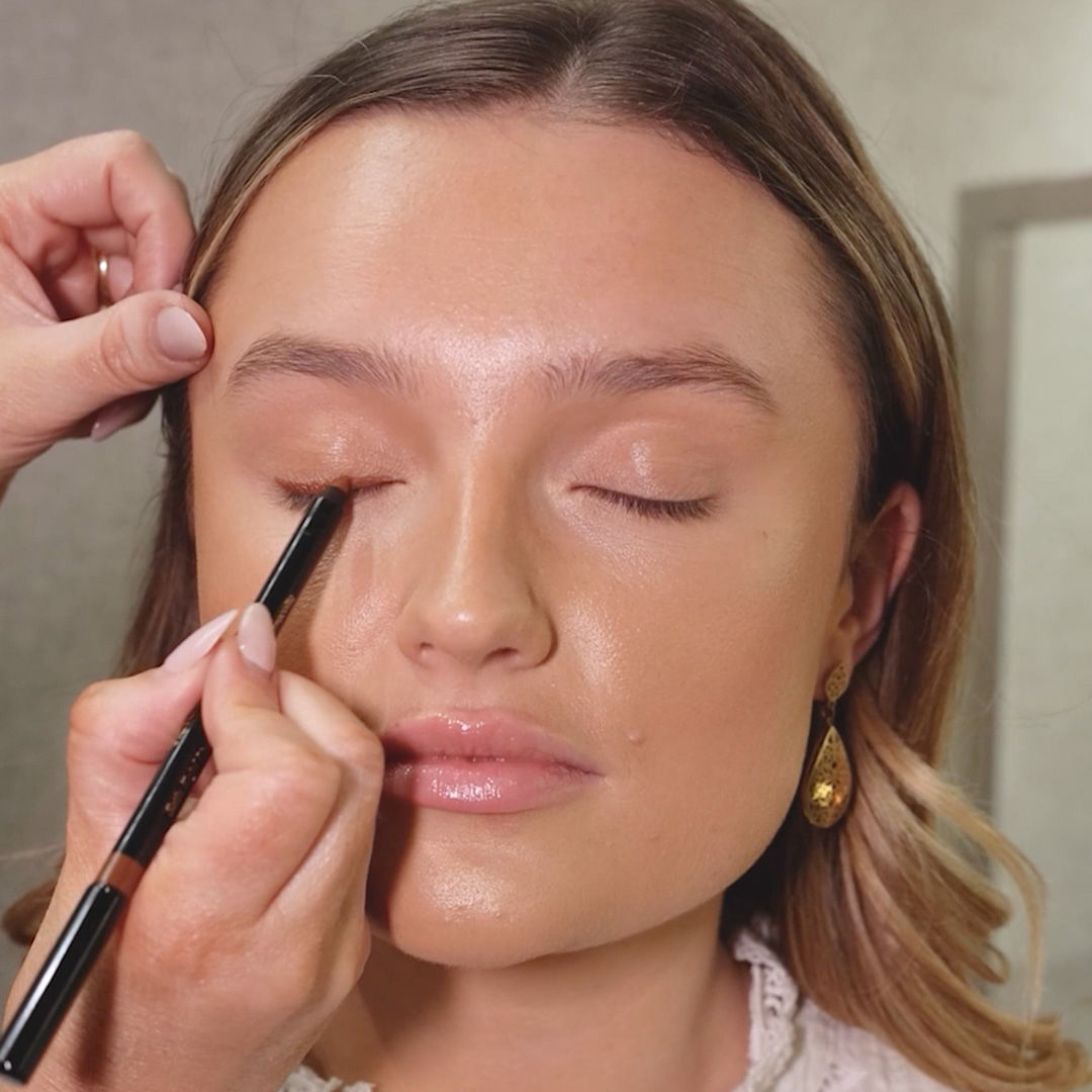 A person getting eyeliner applied to their closed eyelid, with hands holding a pencil.