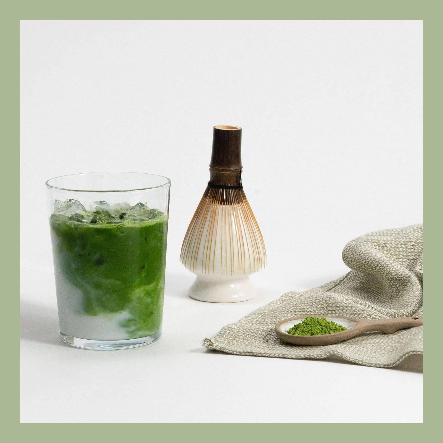 A glass of iced matcha latte, a bamboo whisk, and a spoon with matcha powder on a cloth.
