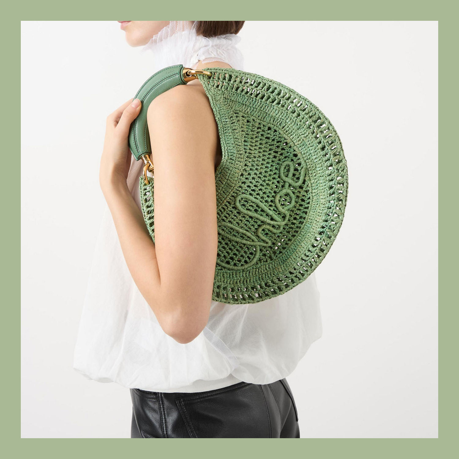 Person holding a green, round, woven bag with 'Chloé' embroidered on it, wearing a white top and black bottoms.