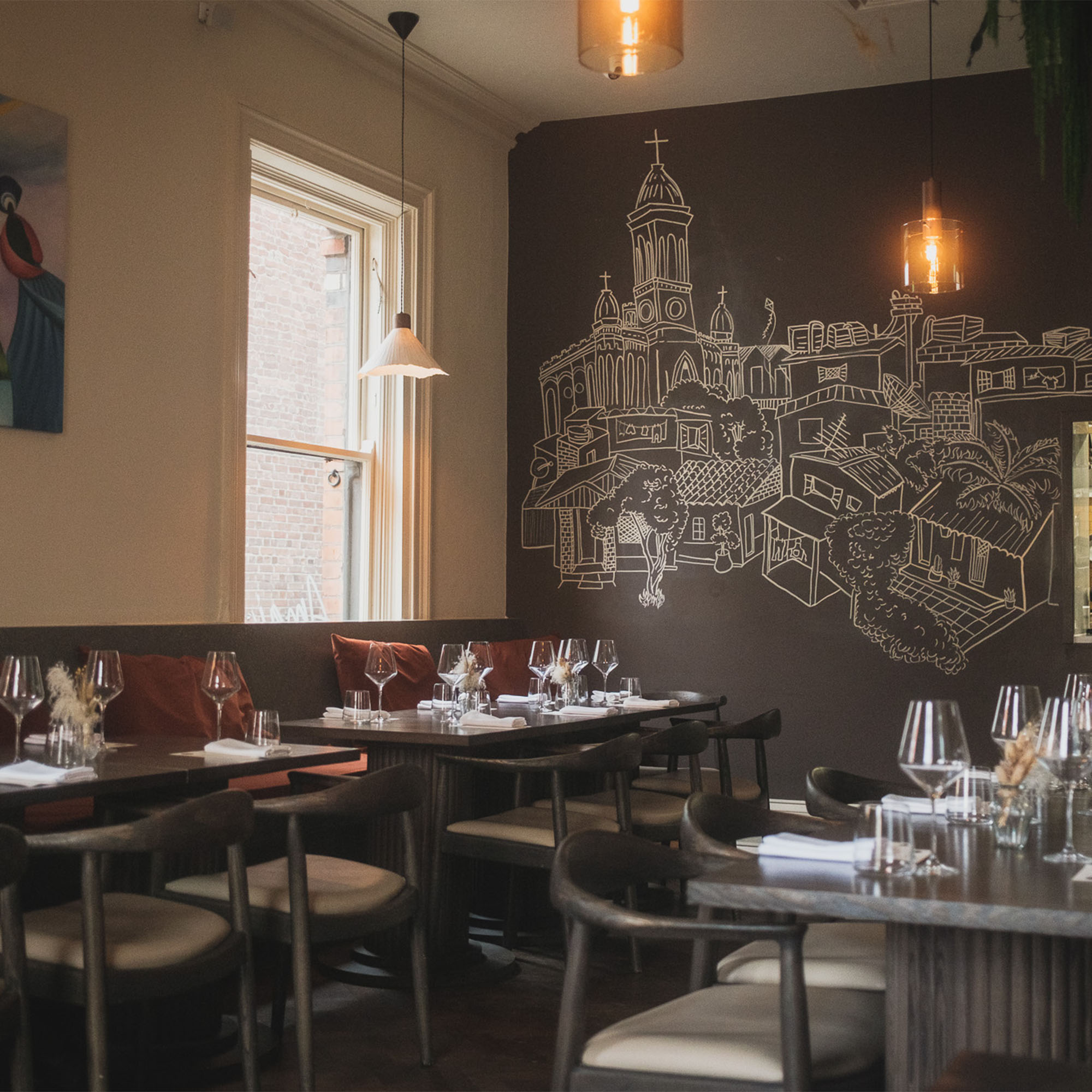 Cozy restaurant interior with tables set for dining. A chalk mural of a church and cityscape decorates the wall, lit by warm hanging lights.
