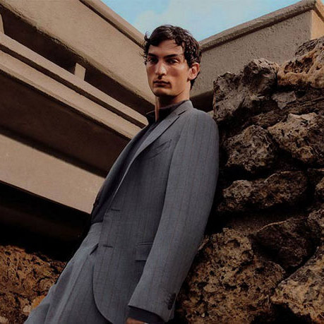 A man in a gray suit poses outdoors near large rocks and a concrete structure.