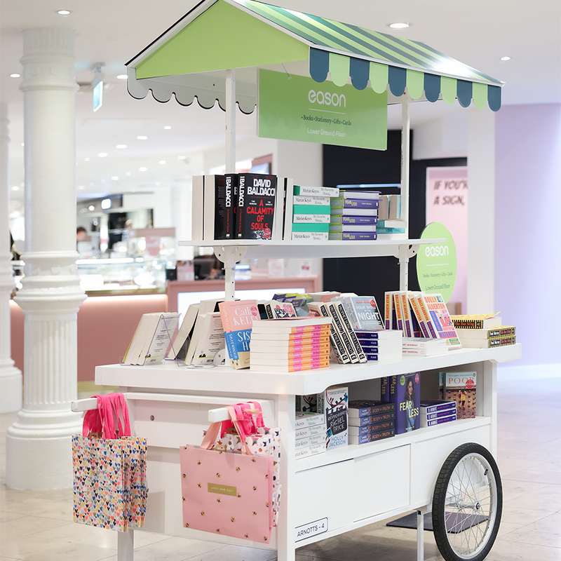 A mobile book cart with shelves of colorful books and fabric bags on display in a well-lit indoor area.