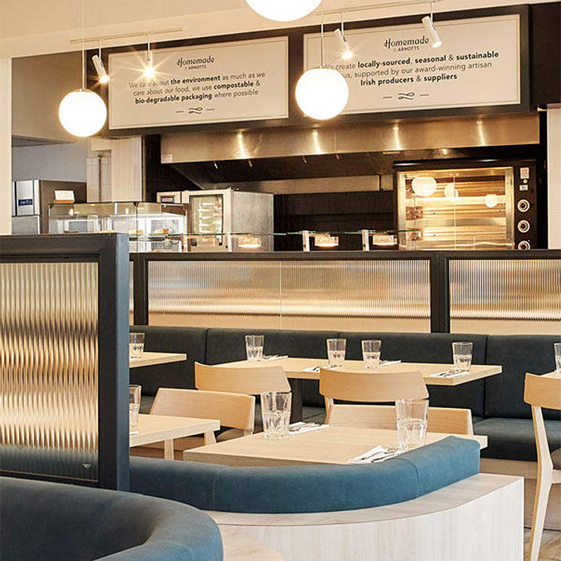Modern restaurant interior with wooden tables, blue chairs, and pendant lights near an open kitchen.