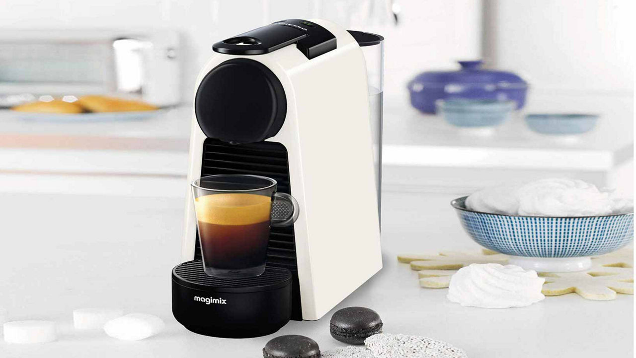 Black and white coffee maker sitting on a white bench in a bright kitchen. A coffee cup full of coffee sits in the machine and coffee pods sit on the counter beside the machine.